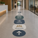 A series of oval and rounded rectangular floor decals leading down a modern office hallway. The decals feature icons and text for "RECEPTION," "VISITORS CHECK-IN," "MEETING ROOMS," and "ELEVATOR" in a coordinated blue, green, and cream color palette.