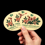 A hand holding three overlapping oval stickers featuring the "Liberty Botanicals" logo with a vintage floral illustration on a cream background.