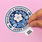 Round wild blooms and botanicals peel tab sticker being peeled open by hand on a pink surface.