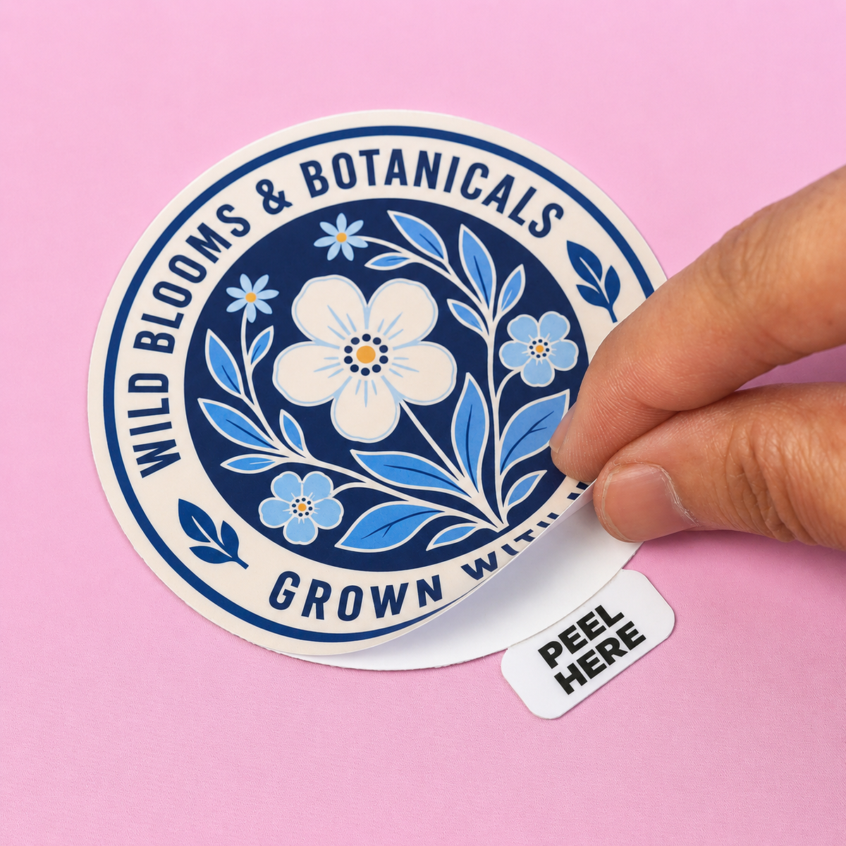 Round wild blooms and botanicals peel tab sticker being peeled open by hand on a pink surface.