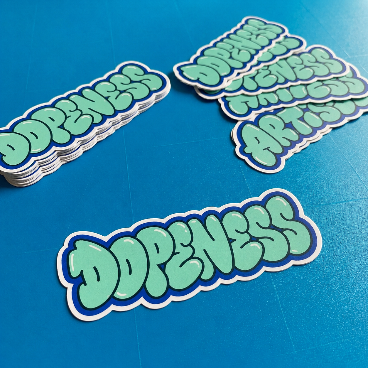 A stack of die-cut premium vinyl stickers featuring "Dopeness" in mint green bubble graffiti letters with a thick blue border, displayed on a vibrant blue background.