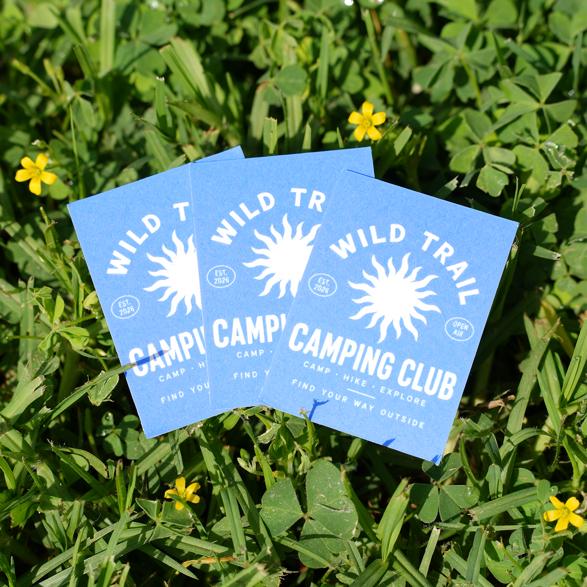 Three blue rectangle stickers featuring a 'Wild Trail Camping Club' design with a sun graphic and text, placed on green grass