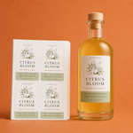Premium rectangle vinyl product labels printed on sheets shown next to a clear glass bottle filled with botanical syrup on an orange background.