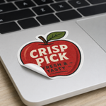 A die-cut removable vinyl sticker featuring a red apple with the text Crisp Pick applied to a silver laptop, with one edge peeling back to show it is easily removable.