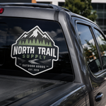 A rugged outdoor-themed car sticker on a truck's rear glass. The design is shield-shaped with snow-capped mountains, green pine trees, and the text "NORTH TRAIL SUPPLY - OUTDOOR GOODS - EST. 2019" in bold white and green typography.