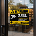 Yellow and black permanent security warning sticker applied to a glass entrance door, stating 24-hour video surveillance and authorized personnel only.