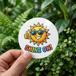A hand holding a custom waterproof sticker featuring a smiling sun with sunglasses in a lush green garden setting.