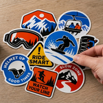 Hand holding collection of die cut snowboard stickers featuring safety warning and snow sport themed designs