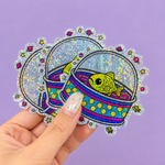 Hand holding fishbowl glitter stickers with a bright space-themed design on a purple background.