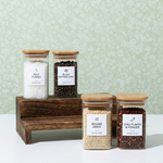 Four glass jars with wooden lids containing different spices on a white surface with a light green patterned wall background.