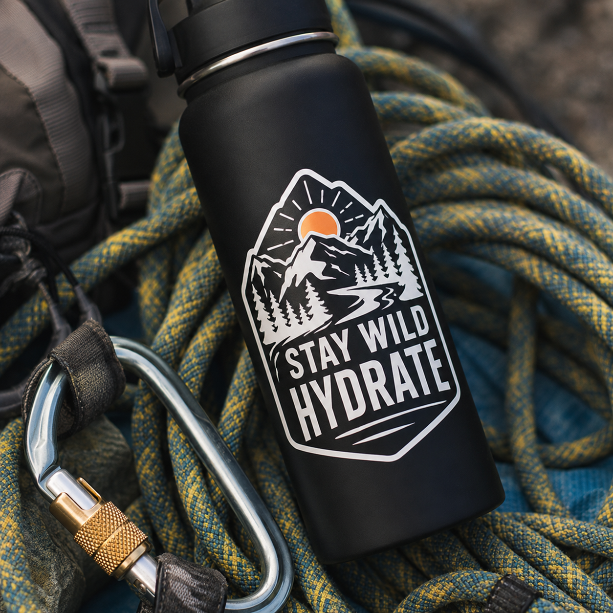 A custom die-cut vinyl decal with mountain scenery and "Stay Wild Hydrate" text on a black insulated water bottle, surrounded by climbing ropes.