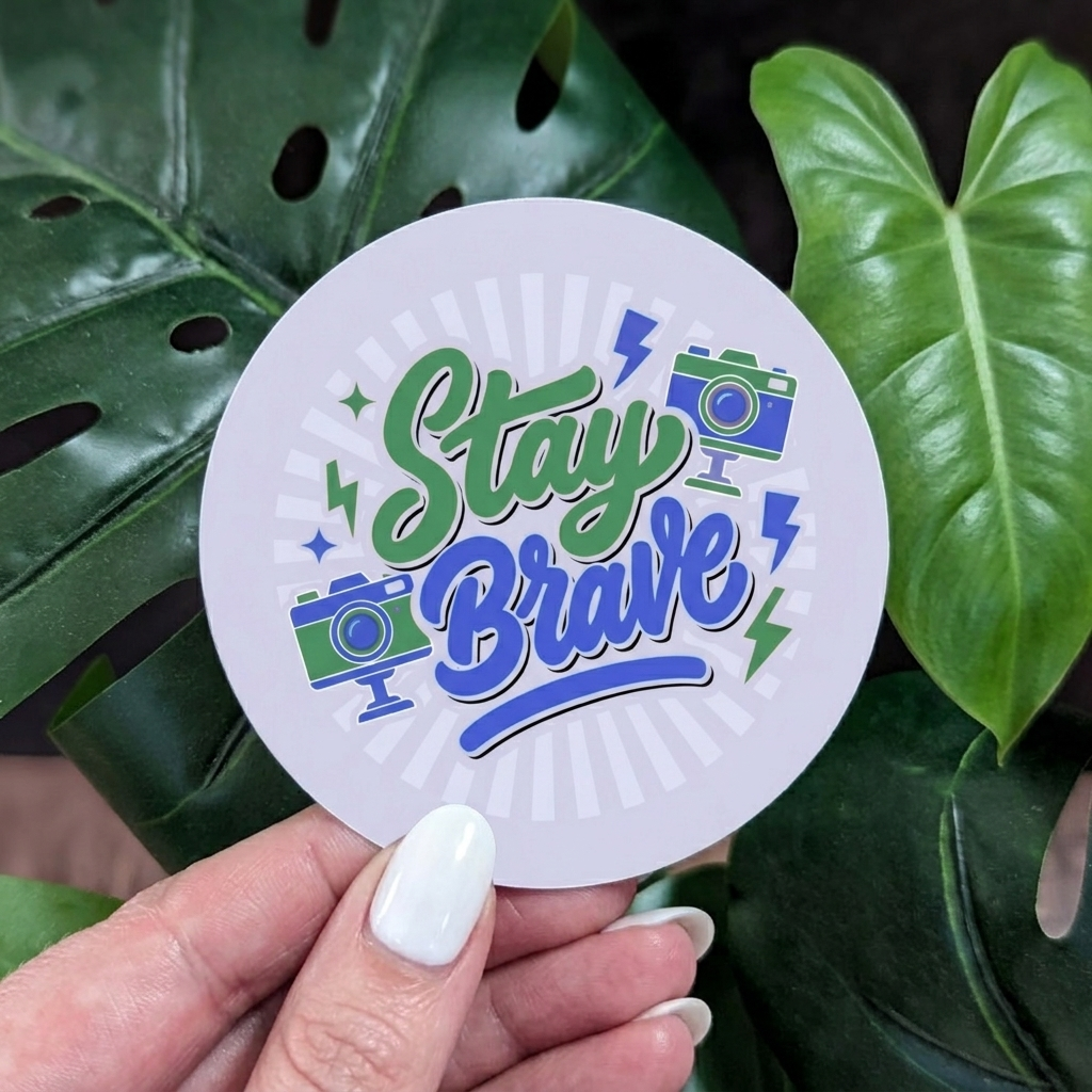 Circular vinyl sticker with 'Stay Brave' text held over green leaves
