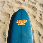 Blue surfboard placed on the sand with a "SURFING NOW!" sticker on the beach background.