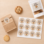 A sheet of round 'Sweet Crumble' chocolate chip cookie stickers next to a brown paper bag and a white pouch, showing custom food packaging label applications.