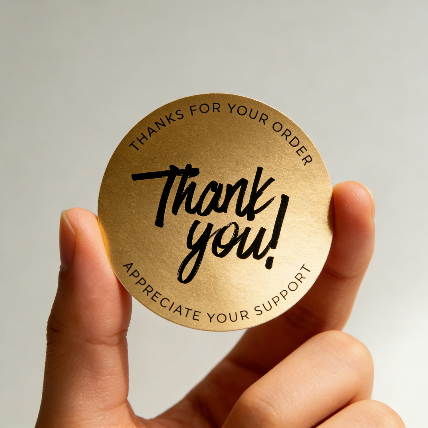 Golden circular sticker with 'Thank you!' text held by a hand against a neutral background