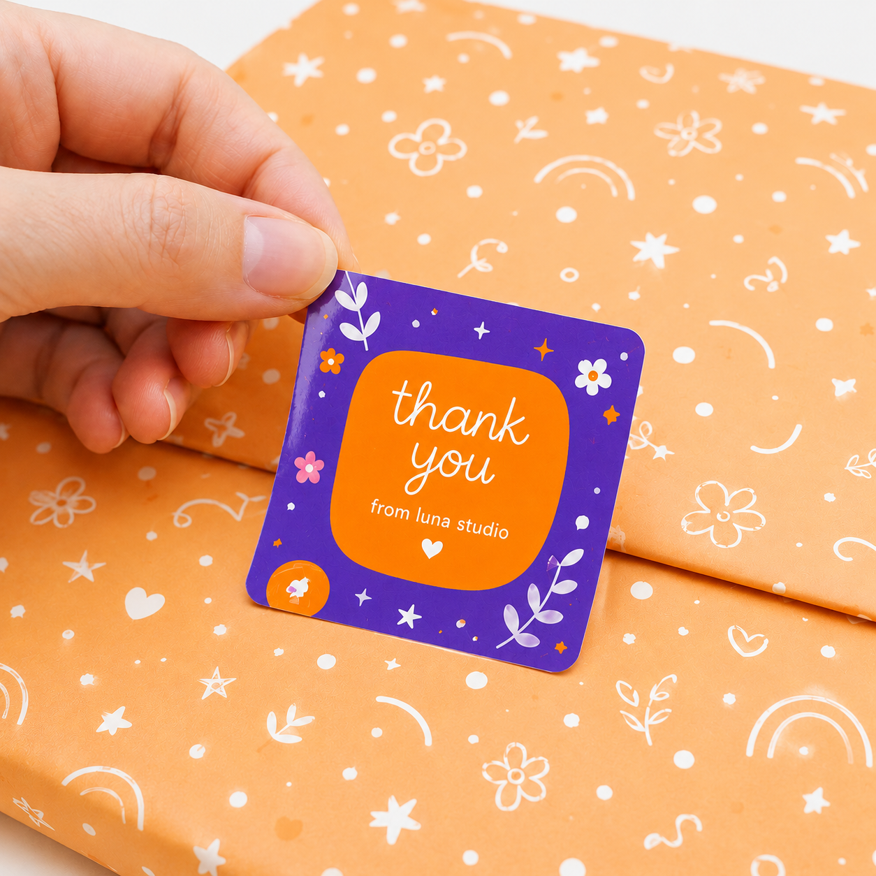 Close-up of glossy custom thank you sticker in purple and orange design, held over orange gift wrap with minimal doodle pattern, premium packaging aesthetic.