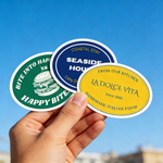 Three custom oval waterproof vinyl stickers (Happy Bites, Coastal Stay, La Dolce Vita) held in hand against blue sky, ideal for water bottles, laptops, and small business branding.