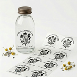 Custom transparent labels with black botanical illustrations applied to a glass bottle and displayed on a roll of sticker sheets.