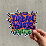 A hand holding a sharp, multi-layered die-cut sticker that reads "Urban Kings Collective" in bold purple and orange graffiti script with a halo detail.