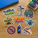 A diverse collection of street art stickers including spray cans, a "RIOT" wordmark, a gas mask, and an octopus, arranged on a wooden desk next to professional markers.