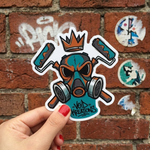 A high-quality vinyl graffiti sticker of a stylized teal and orange gas mask with paint rollers and a crown, held against a red brick wall background with urban tags.
