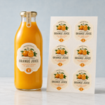 Custom wavy-edge vinyl label sheets next to a glass orange juice bottle, demonstrating oil-resistant and waterproof vinyl sticker quality for beverage packaging.
