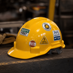 A yellow construction hard hat sitting on a workbench, detailed with multiple safety stickers like Safety First, Head Protection, PPE Required, and Watch Your Step.
